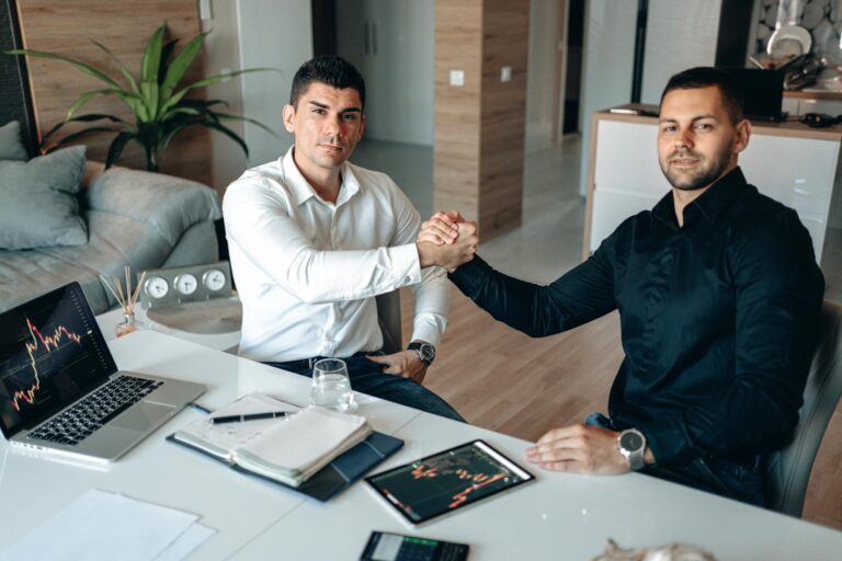 Two businessmen shaking hands at a modern office desk with tech devices, indicating a successful partnership.